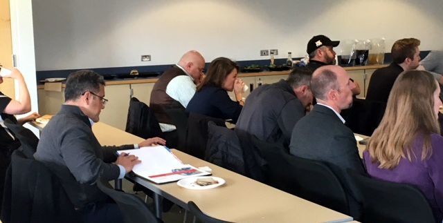 People in a meeting, seated at tables. A man in a suit is looking at papers. Others are listening, some with heads down.