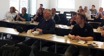 People seated at tables in a classroom setting, listening. Coffee cups and papers on tables.