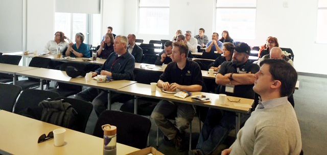 A group of people sitting at desks in a classroom-like setting, possibly listening to a presentation.