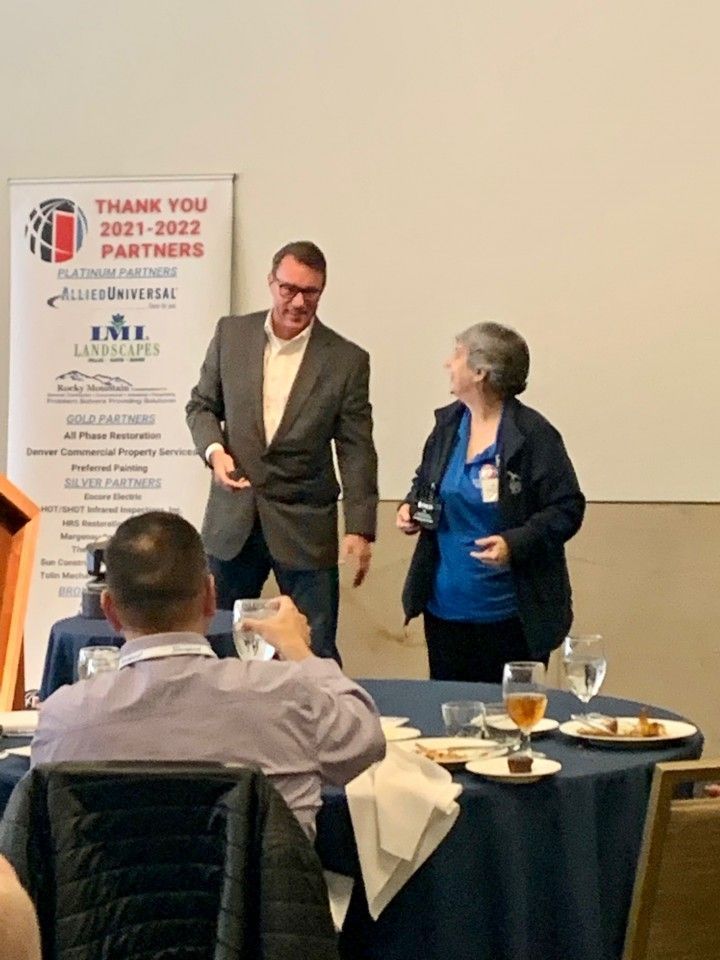 Man in suit and woman in blue shirt conversing at event with partners listed on banner.