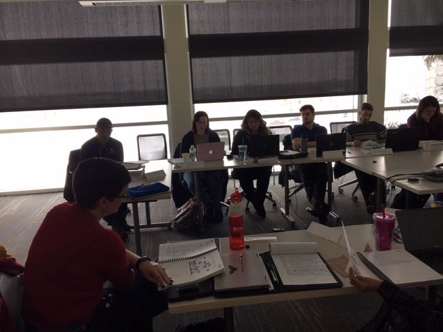 People seated around a table with laptops, participating in a meeting; windows with blinds.