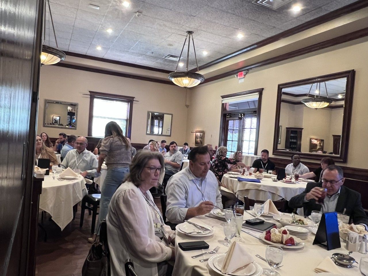 People seated at tables in a restaurant, a server attends. Ornate ceiling lights and mirrors decorate the interior.