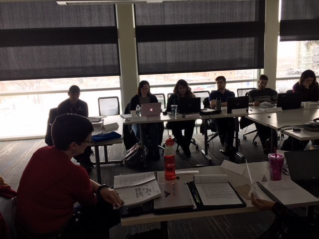 People seated around a long table in a room with a large window.  They appear to be in a meeting.