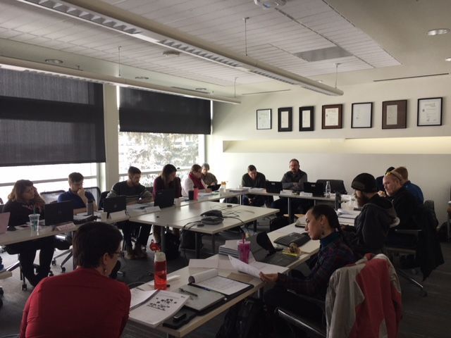 People seated at a large table in a conference room. Papers and laptops are visible. Natural light streams in.