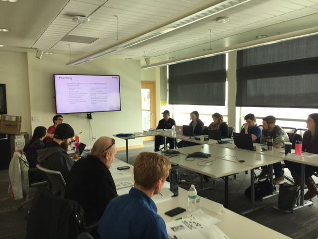 People seated around a table, looking at a screen during a meeting. Bright room with large windows.