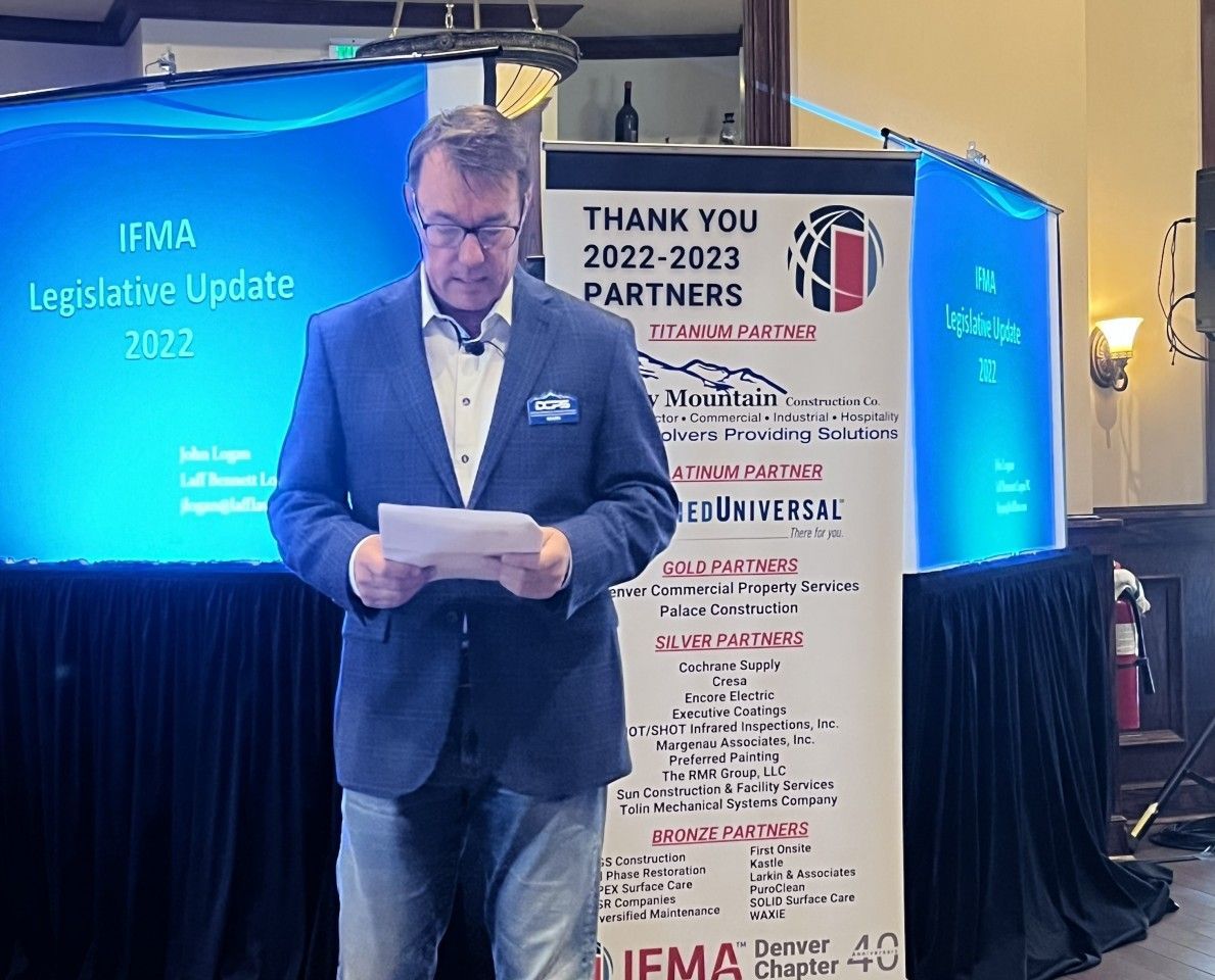 Man in blue blazer reads from papers at podium, event banner behind him.