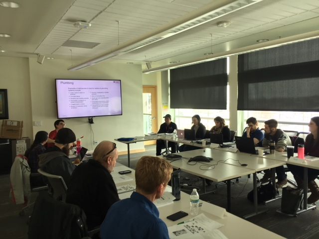 Group of people at a table in a meeting, laptops and presentation screen visible.