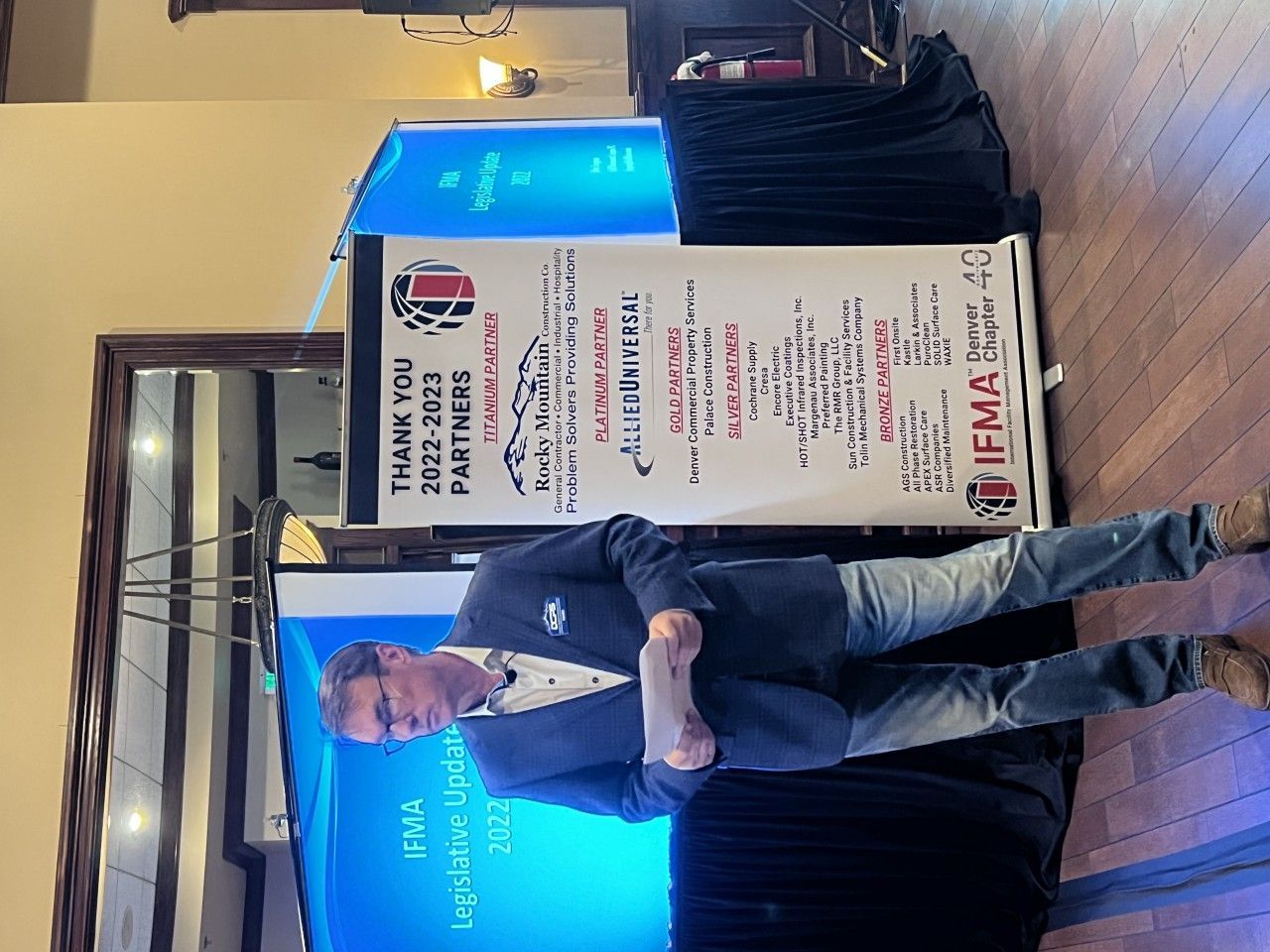 Man in suit stands near banner, looking at the audience, indoors.