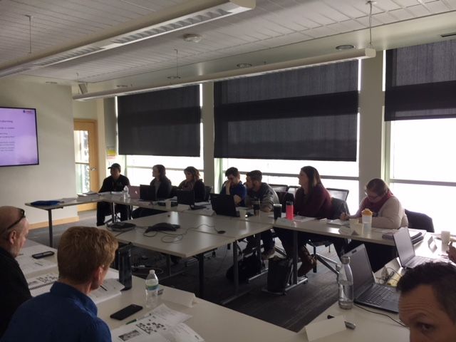 People seated at a table in a meeting room with laptops. Presentation on screen.