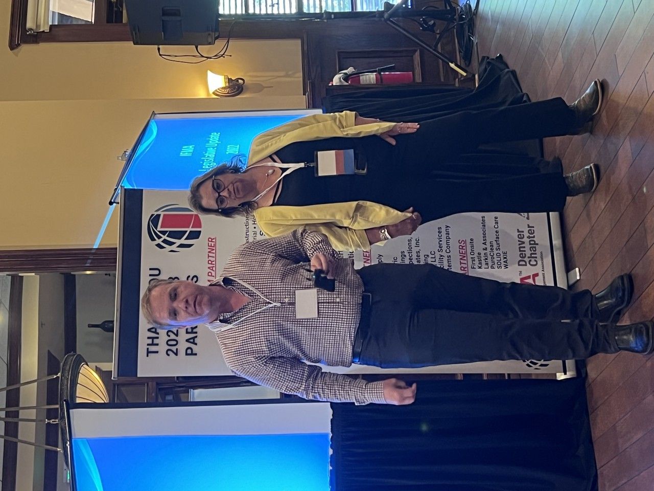 Two people stand near a banner, one holding a microphone. They appear to be at an event or conference.