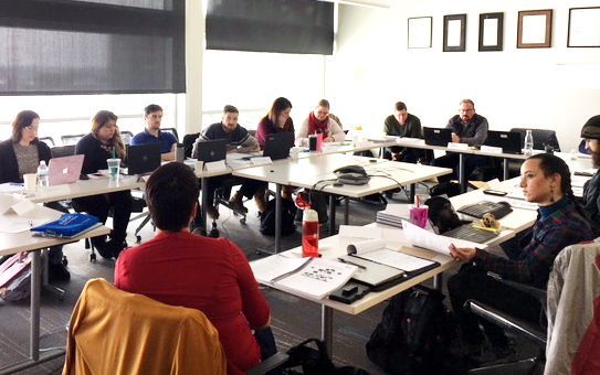 Group of people in a meeting at tables, in a well-lit office. Some use laptops.