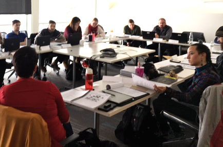 People seated at tables in a meeting. A woman in red speaks, facing others who are using laptops.
