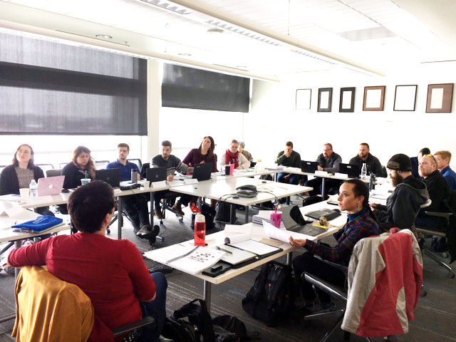 People in a meeting room, seated around tables. Some are looking at papers, laptops, and each other.