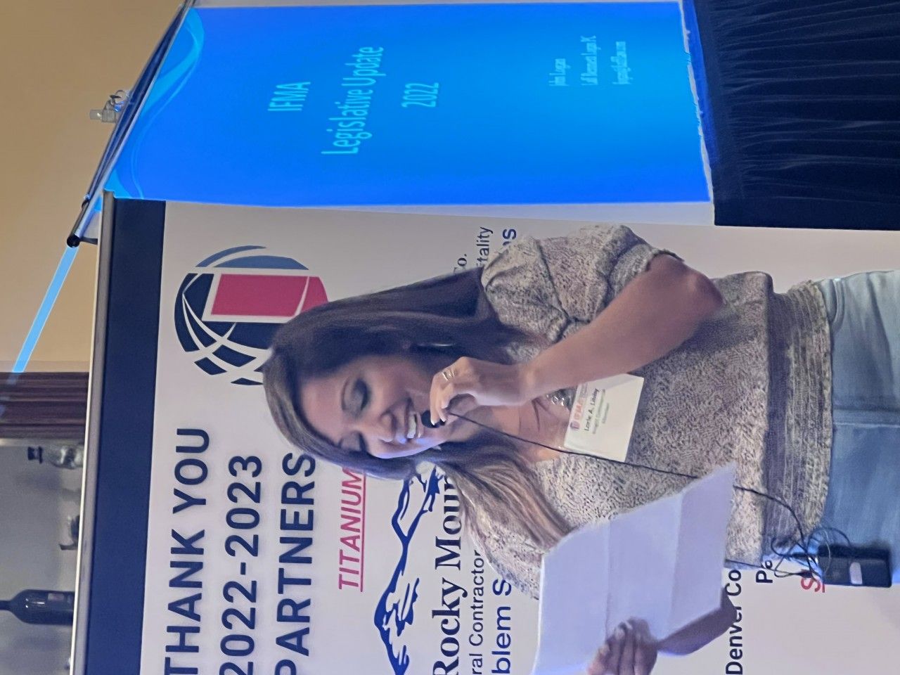 Woman speaking at a conference. She holds papers, smiles, stands near a podium with a banner and screen.