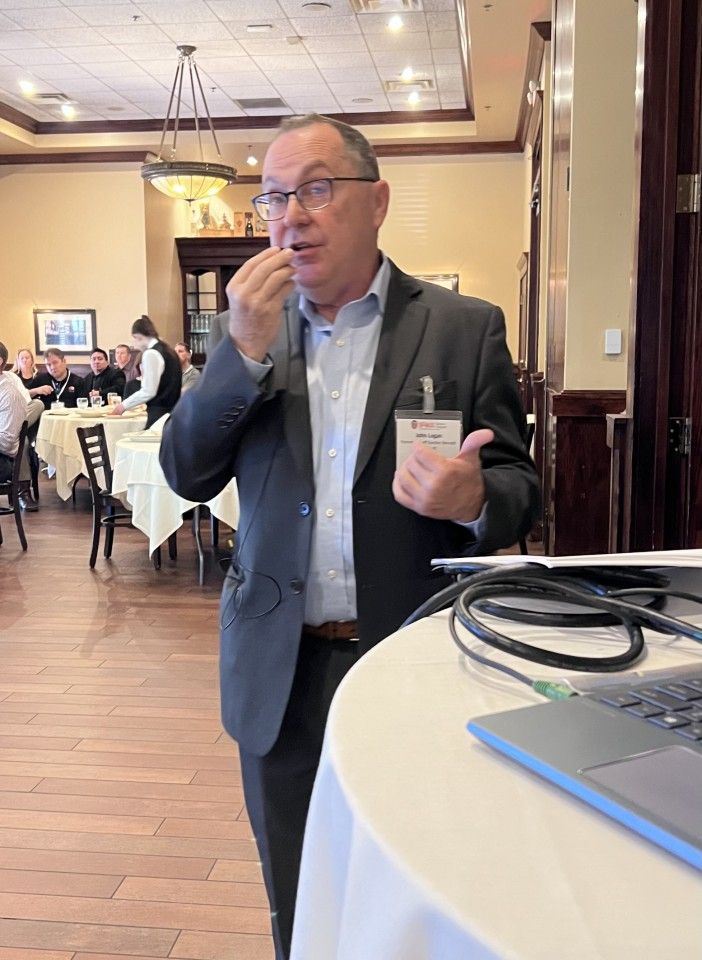 Man in suit speaks at a restaurant. He gestures, holds something near his mouth, and has a name tag.