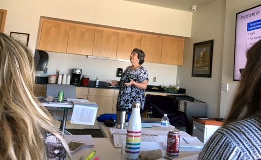 Woman presenting in a room, gesturing towards a screen with text. People seated at a table, some with drinks.