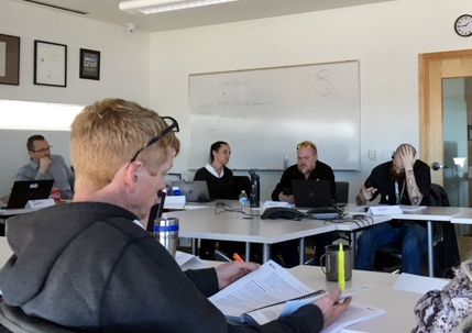 People seated around a table in a meeting room; some are looking at papers, one man is speaking, others have laptops.