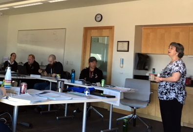 A woman speaks to a group seated at a table in a bright room.