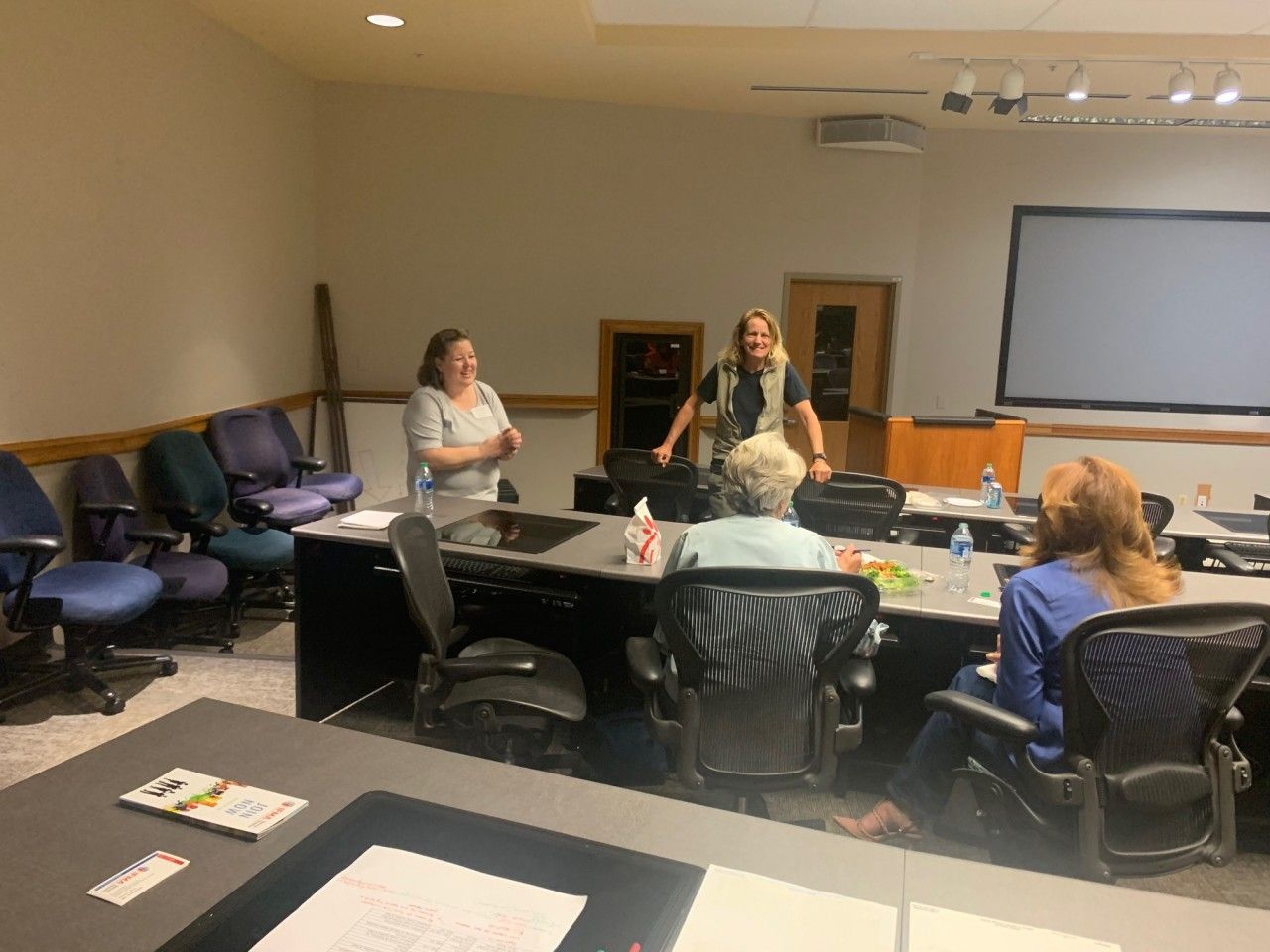 People at a table in a meeting room, two women stand, one speaks, others listen.