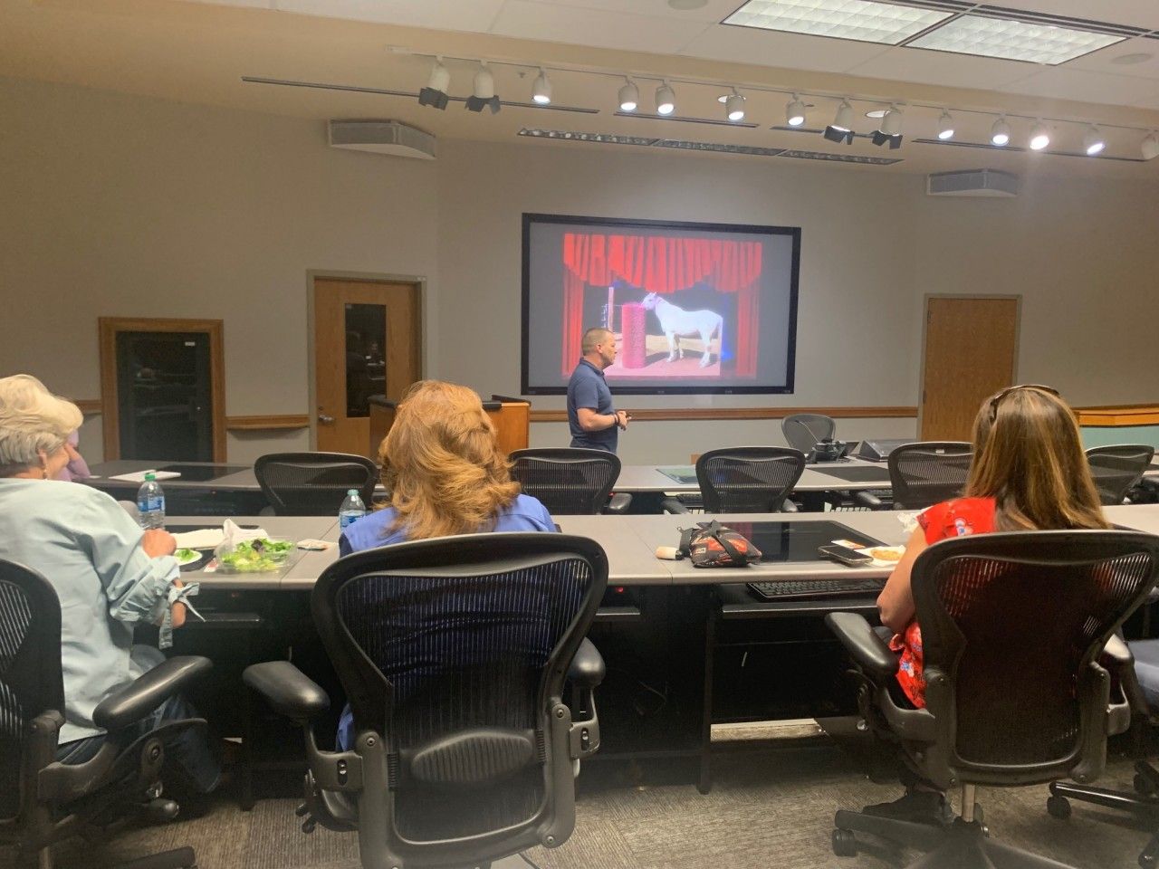 A person giving a presentation with a horse image on a screen in a conference room with three seated attendees.