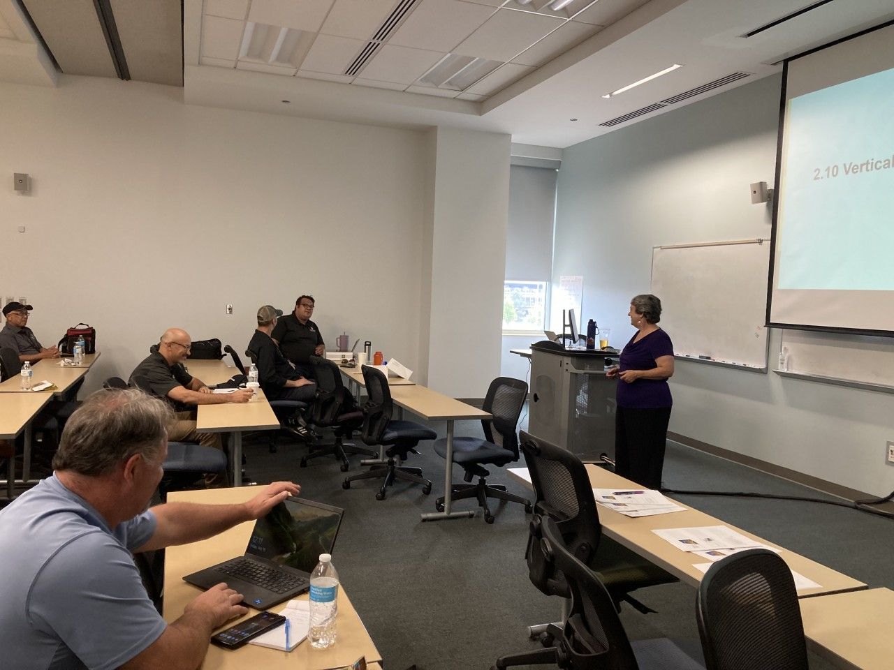 Woman presents in a classroom. Attendees sit at desks, some using laptops. A presentation screen is visible.