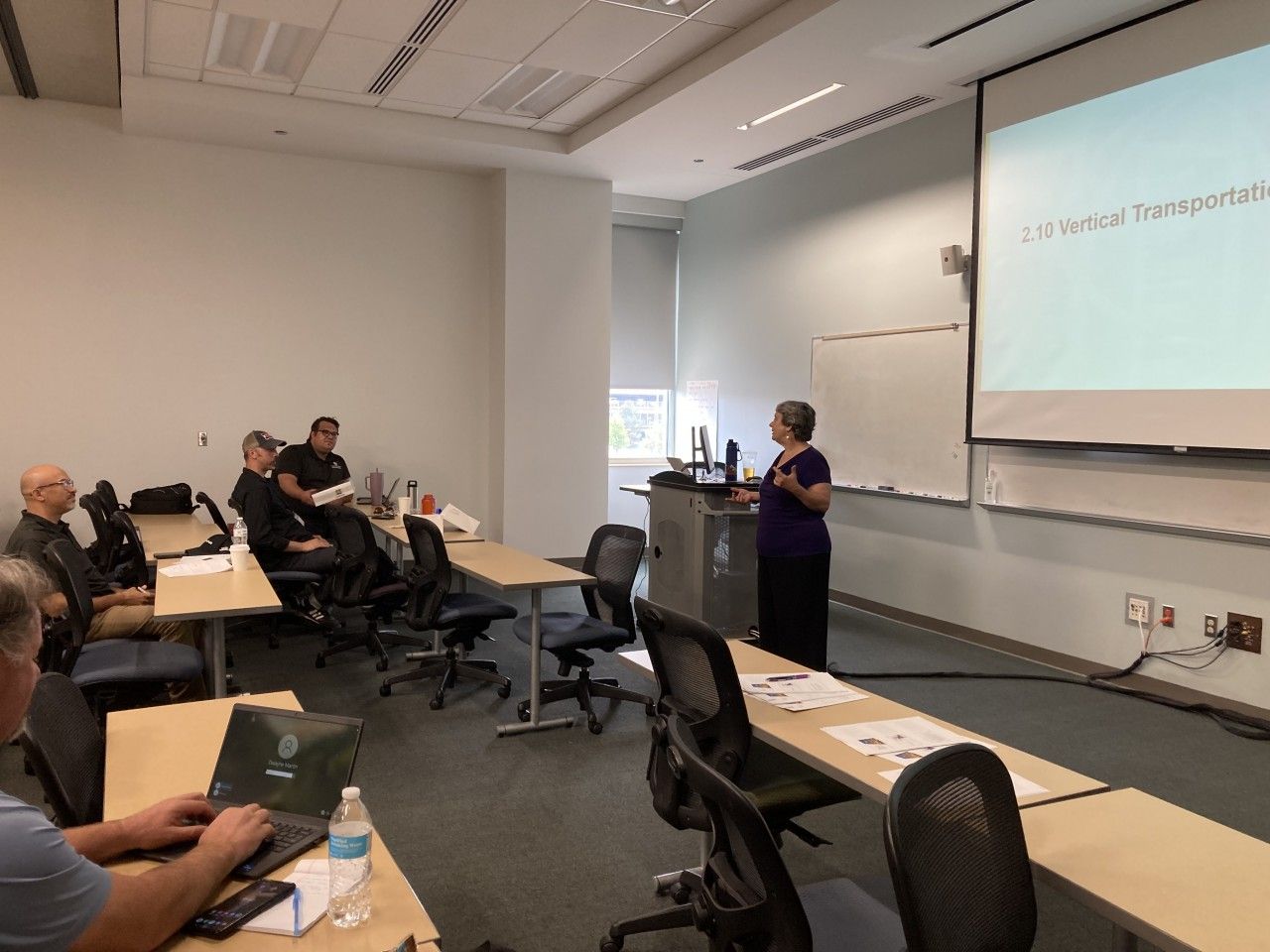 A woman lecturing in a classroom; students seated at tables, one working on a laptop, a projector screen at the front.