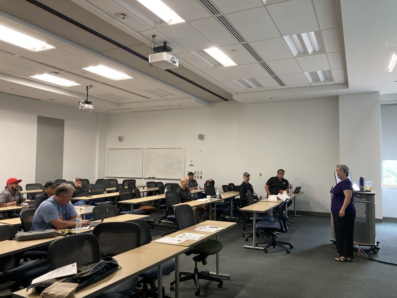 Classroom lecture in progress. People seated at desks, professor at podium, projector on ceiling.