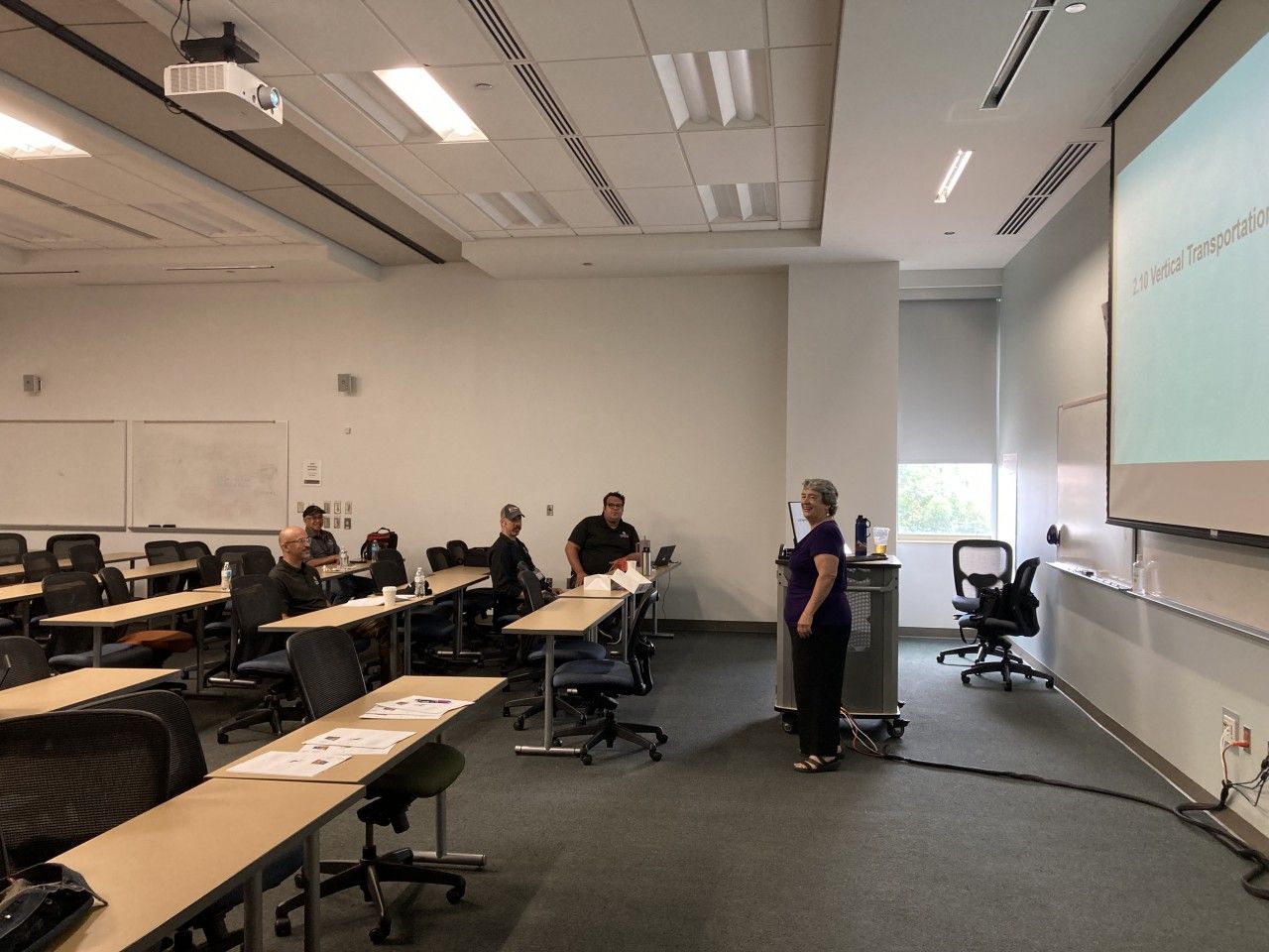 A classroom with a presenter and a few students; a projector screen displays text.