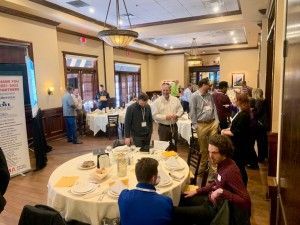 People networking at an event in a dining room; tables set, banner visible.