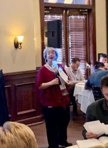 Woman speaking into a microphone at a table; others seated, some taking notes. Interior restaurant setting.