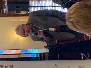 Man in a grey suit speaks into a microphone at a podium, in front of a screen and attendees.