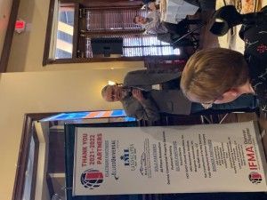 Man speaking at event with banner listing sponsors. Restaurant setting.