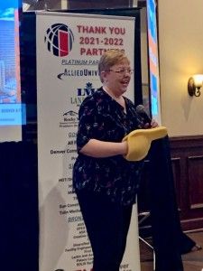 Woman speaking at event, holding a yellow object and dark coat, standing next to a banner.