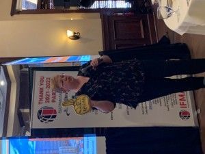 Woman speaking at an event, holding a foam hand. Behind her is a banner that says 