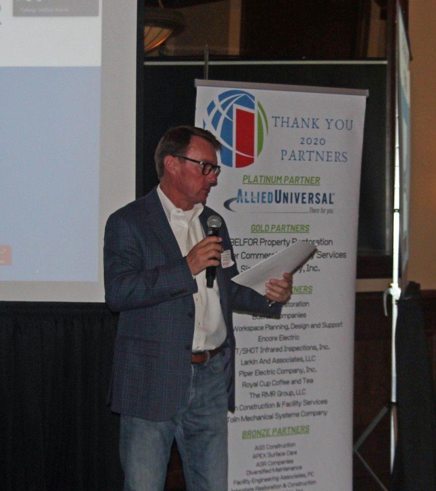 Man in blazer speaks at podium holding paper, banner says