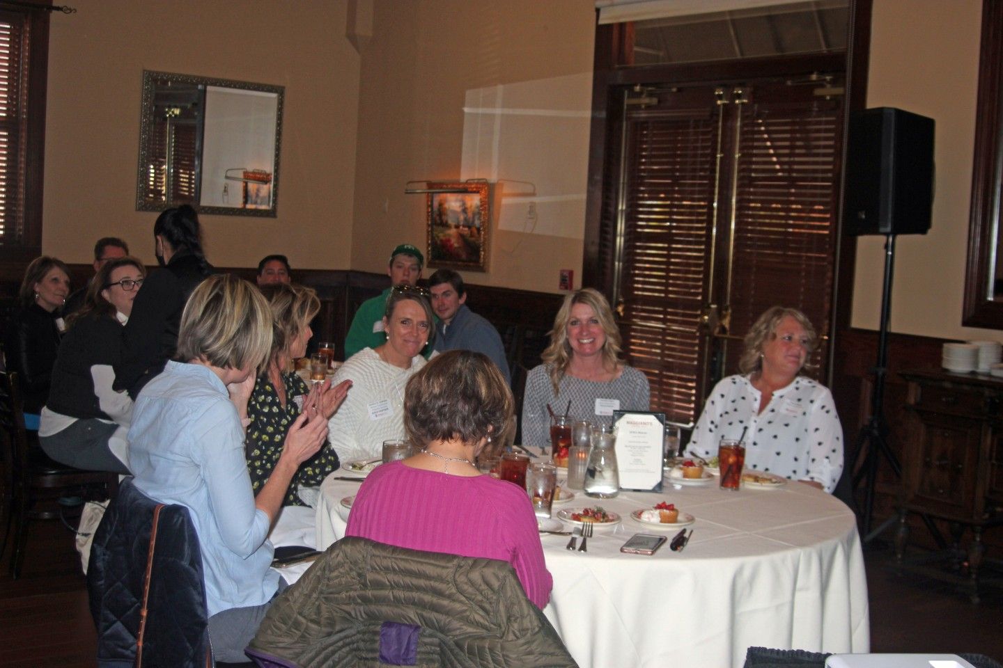 People at a table in a restaurant, a woman applauds, others listen and smile.