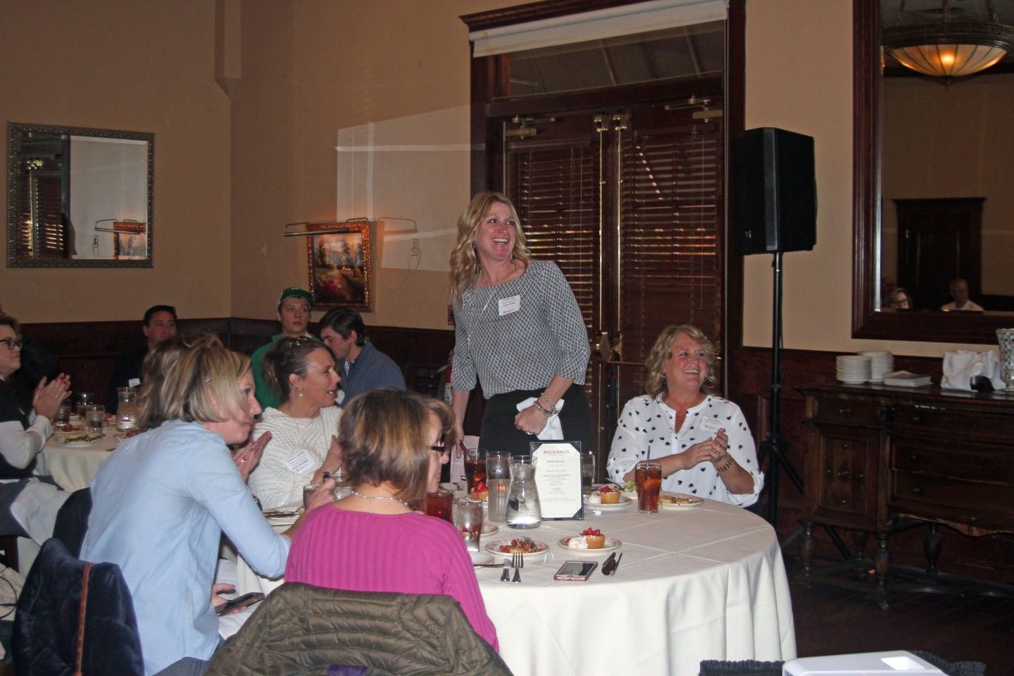 Woman standing, speaking at a table in a restaurant; others seated and listening.