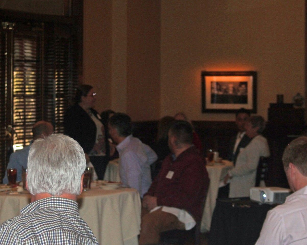 People at tables listen to a woman speaking in a room with a window, art, and a projector.