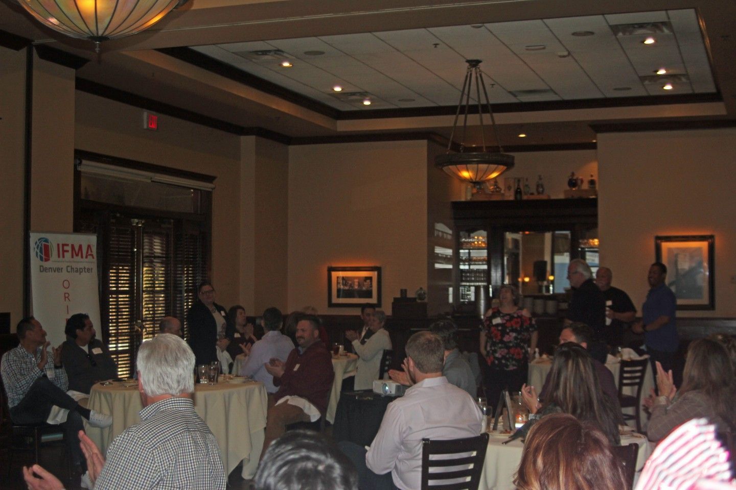 People seated at tables in a restaurant for a meeting; a panel of people is at the front.