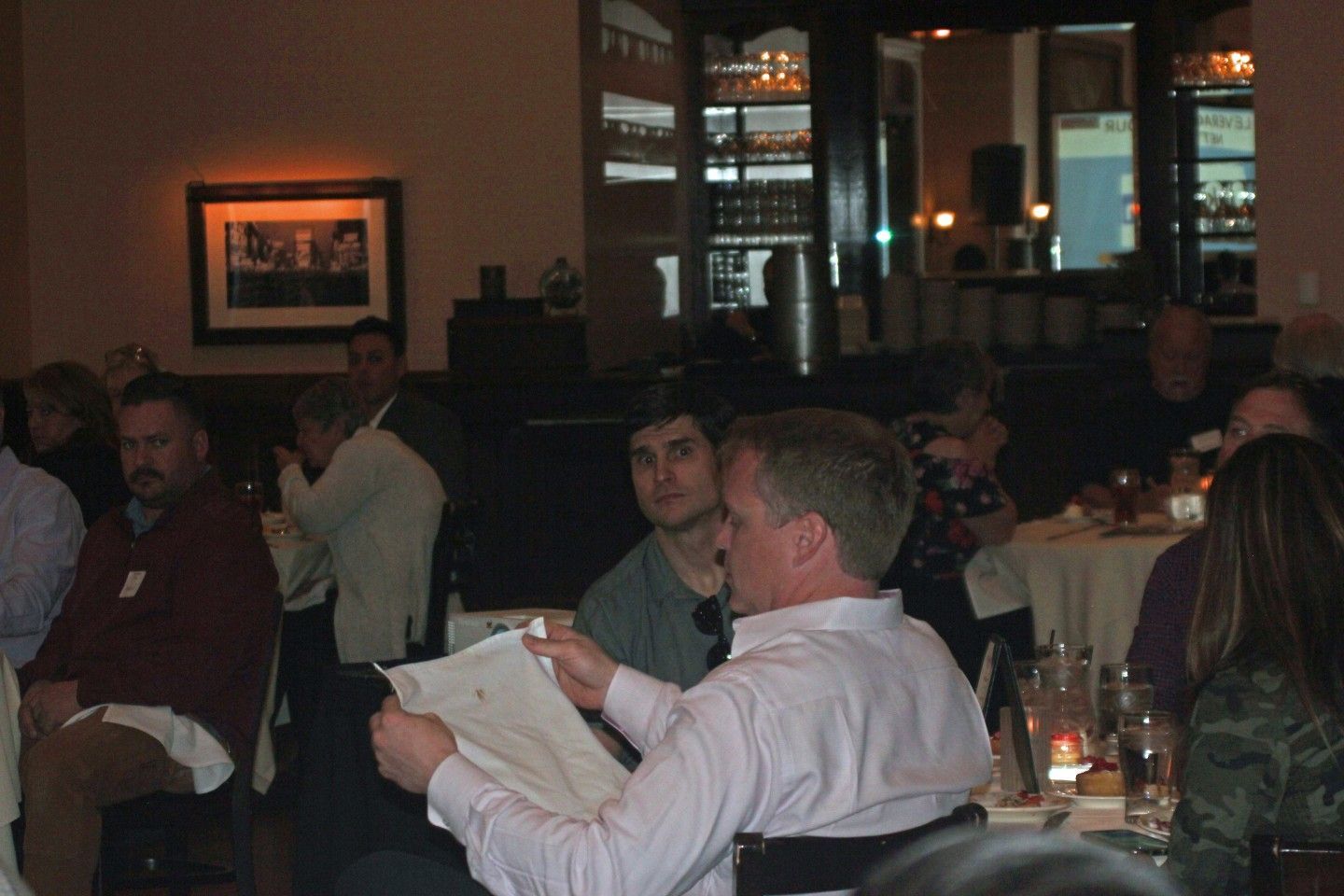 Group in restaurant, man holding paper, others seated, looking on. Soft lighting, neutral tones.