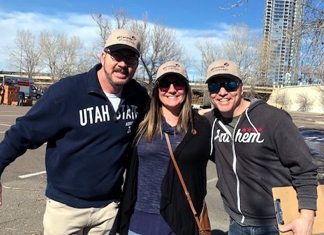 Three people smiling, wearing hats and casual clothing outdoors near a tall building.