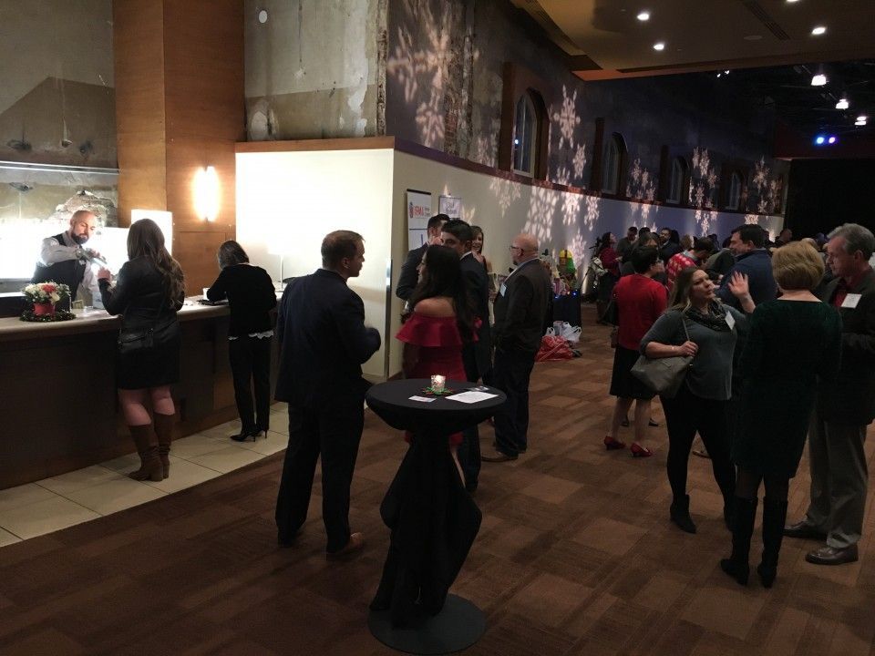 People gathered at an event in a lobby with a bar. Dark walls, wood floor, and various dress attire.