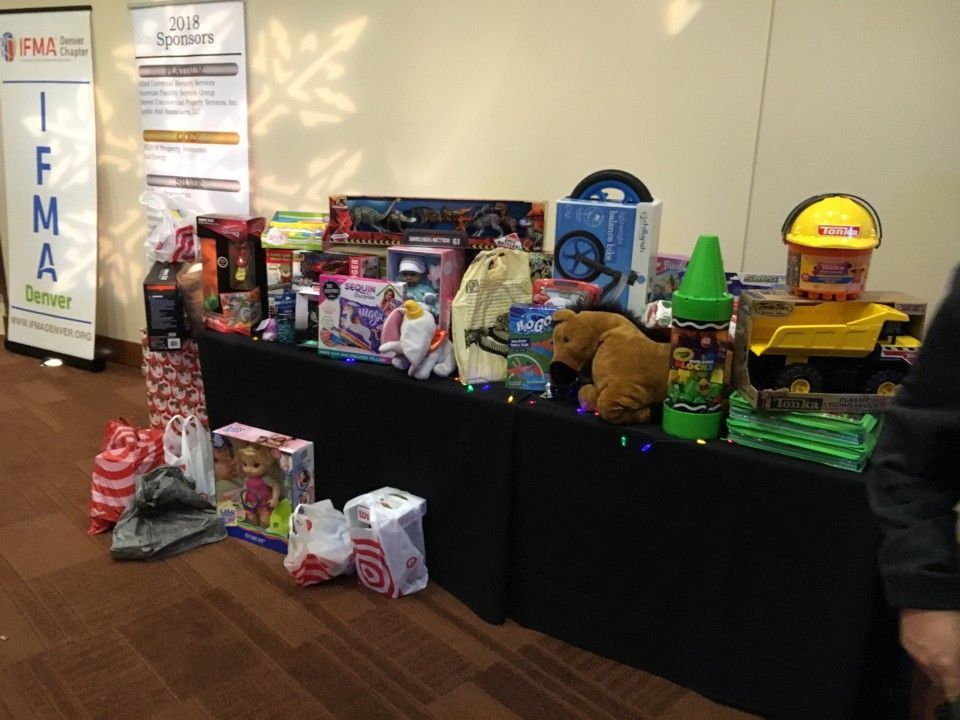 Table of toys and gifts, including a truck, bear, and bags, for a donation.  IFMA banner in the background.