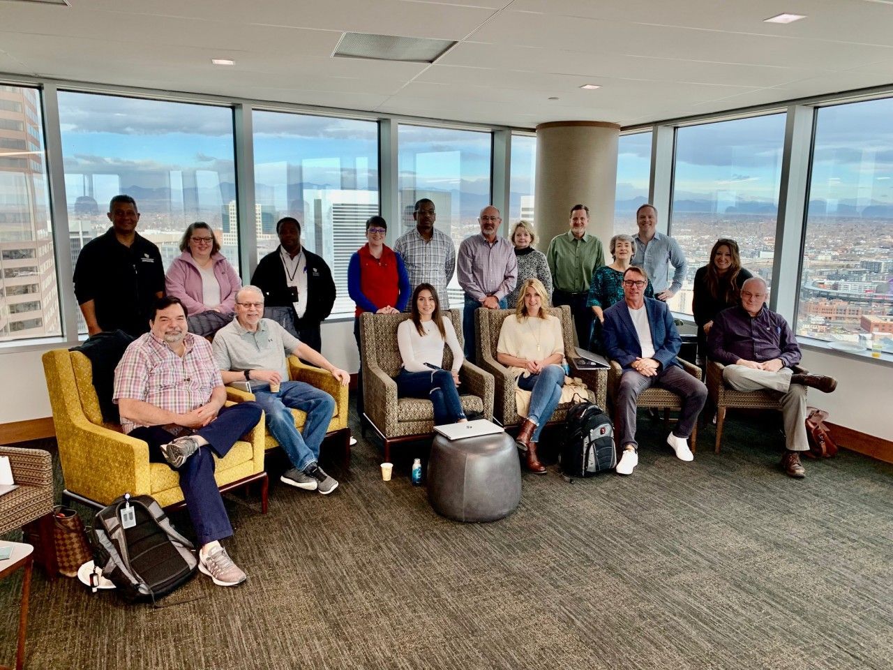 Group of people in a room with large windows overlooking a city, some sitting and some standing.