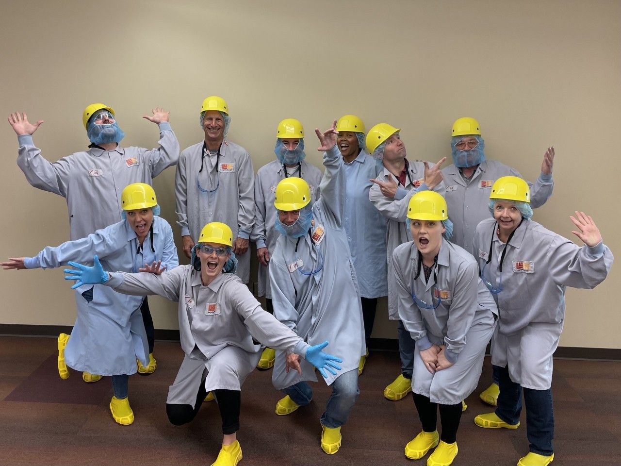 Group of people in yellow hard hats, lab coats, and booties posing playfully.