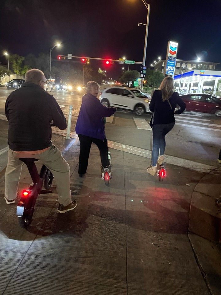 Three people on scooters by a street at night near a gas station and traffic.