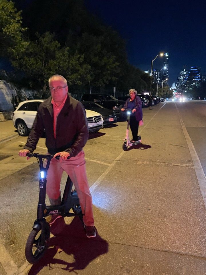 Two people ride scooters on a city street at night.
