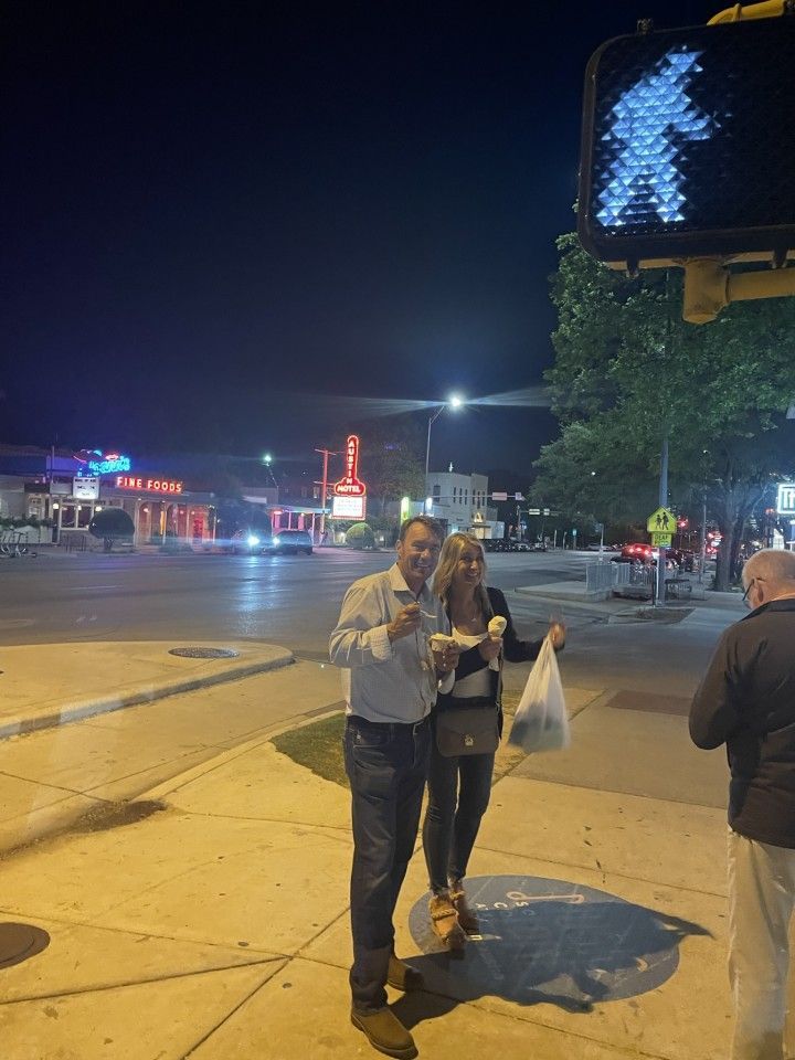 A couple smiling and holding ice cream on a sidewalk at night, near a crosswalk signal.