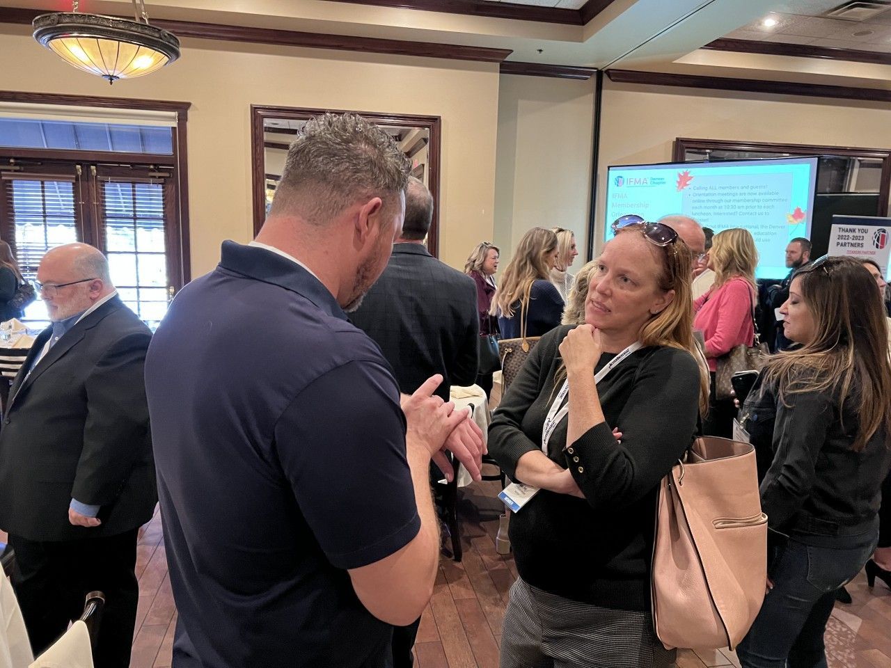 People networking at an event. Man in dark shirt conversing with woman in black, others nearby. Indoor setting.