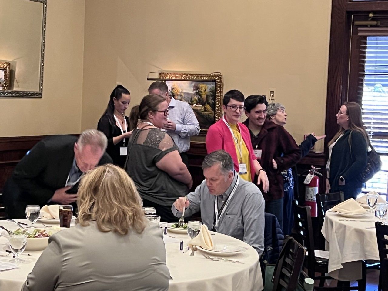 People at a restaurant, some standing near tables, others eating. Soft lighting and a neutral color scheme.